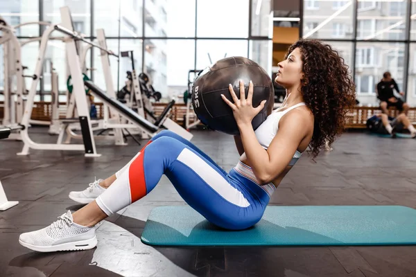 Štíhlá černovlasá dívka oblečená do sportovních šatů cvičením pro tisk na rohoži pro fitness a fitness Ball v tělocvičně — Stock fotografie