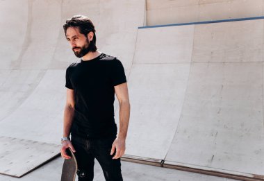 Elinde kaykay tutan şık rahat giysiler giymiş adam güneşli bir günde slayt yanında bir paten parkı duruyor