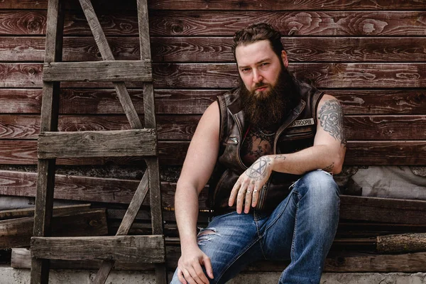 Forte homem brutal com uma barba e tatuagens em suas mãos vestida com colete de couro e jeans senta-se em um fundo de parede de madeira ao lado de uma escada de madeira fora — Fotografia de Stock
