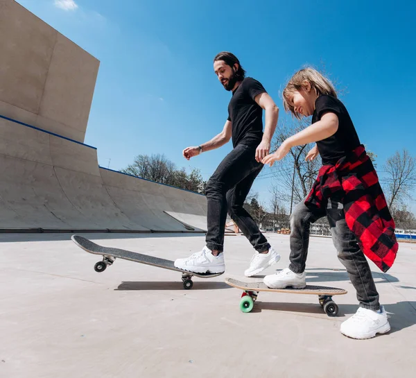 Father and his little son dressed in the casual clothes ride skateboards in a skate park with slides at the sunny day