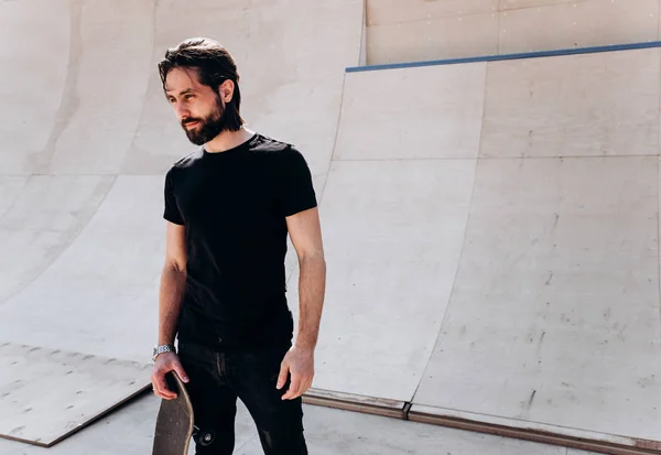 Der Mann in der stylischen Freizeitkleidung mit dem Skateboard in der Hand steht an einem sonnigen Tag in einem Skatepark neben der Rutsche — Stockfoto