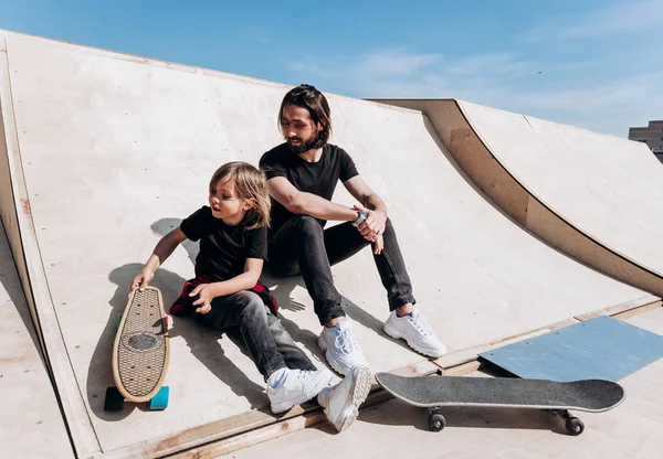 The young father and his son dressed in the stylish casual clothes are sitting together on the slide next to the skateboards in a skate park at the sunny warm day