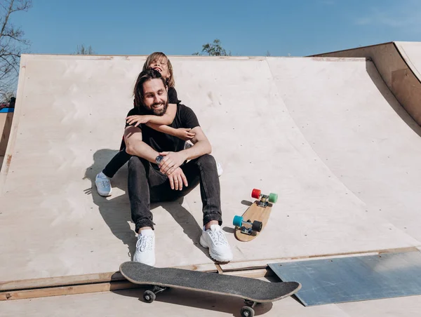 Happy young father and his son dressed in the stylish casual clothes are sitting in an embrace together on the slide next to the skateboards in a skate park at the sunny warm day