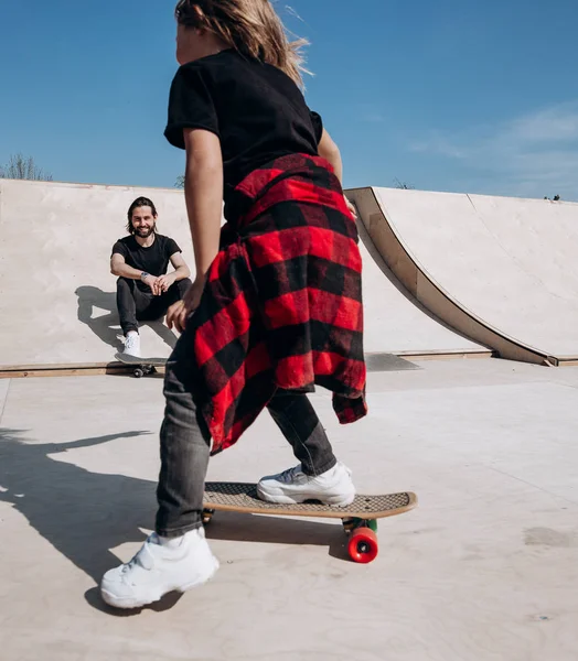 Father and his little son dressed in the casual clothes ride skateboards and have fun in a skate park with slides outside at the sunny day