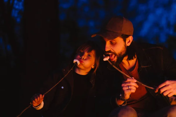 Feliz padre y su pequeño hijo sentados en los troncos del bosque frente a un fuego y comiendo malvaviscos de las ramitas — Foto de Stock