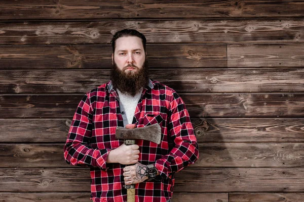 Жорстокий міцний чоловік з бородою, одягнений у перевірену сорочку і рвані джинси, стоїть з сокирою в руках на тлі дерев'яної стіни — стокове фото