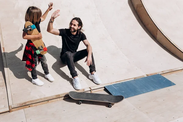 The young father and his son dressed in the stylish casual clothes are sitting and have fun together on the slide next to the skateboards in a skate park at the sunny warm day
