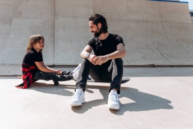 Genç baba ve oğlu günlük kıyafetlerini giymiş güneşli bir günde kaykay parkında oturuyorlar.
