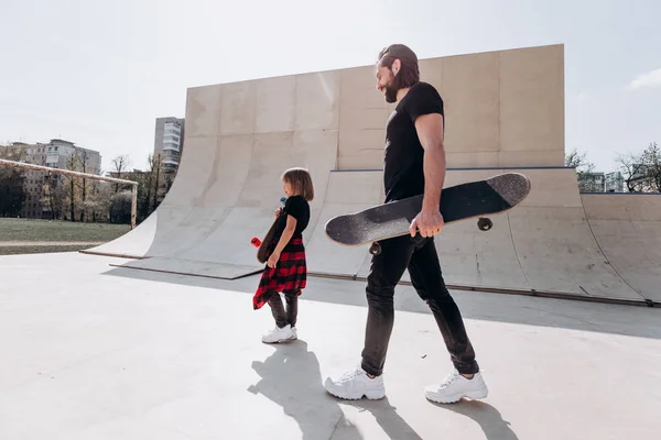 Vader en zijn kleine zoon gekleed in de stijlvolle casual kleren lopen met de Skateboards in hun handen in een skatepark op de zonnige dag — Stockfoto