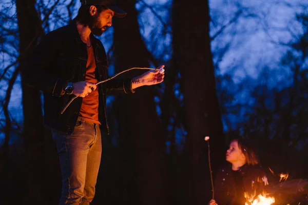 Felice padre sta di fronte a un fuoco nella foresta tenendo rametti in mano la notte . — Foto Stock