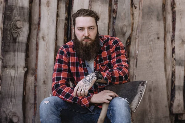 Brutal hombre fuerte con una barba vestida con una camisa a cuadros y vaqueros rotos está sentado con un hacha en las manos contra el fondo de una valla de madera — Foto de Stock