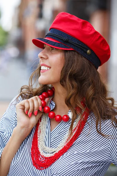 Menina morena elegante vestida com uma blusa listrada, contas vermelhas e um boné vermelho posa na rua da cidade em um dia ensolarado — Fotografia de Stock