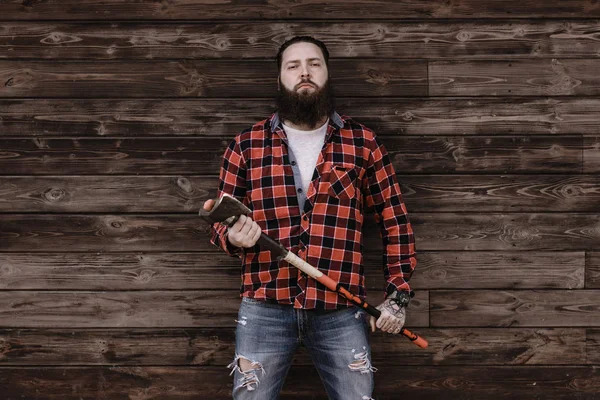 Brute sterke man met een baard gekleed in een geruite overhemd en gescheurde jeans staat met een bijl in de handen op de achtergrond van een houten muur — Stockfoto