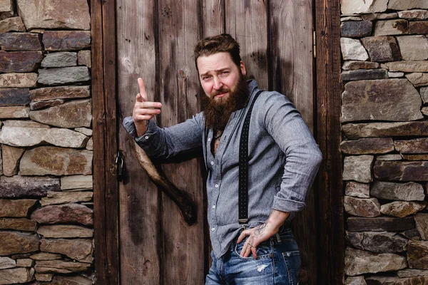 Homme brutal fort avec une barbe et des tatouages sur les mains habillé de vêtements décontractés élégants pose sur le fond de mur de pierre et porte en bois — Photo
