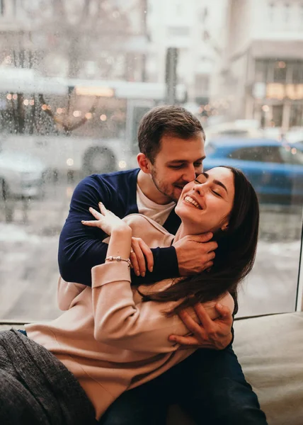 Chico cariñoso abraza a su hermosa novia sentada en el alféizar de la ventana en un acogedor café . —  Fotos de Stock