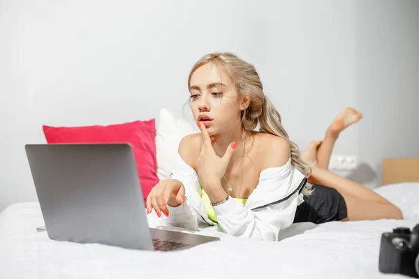 Молода дівчина блогер одягнений в модний одяг лежить на ліжку поруч з камерою і працює в ноутбуці в кімнаті — стокове фото