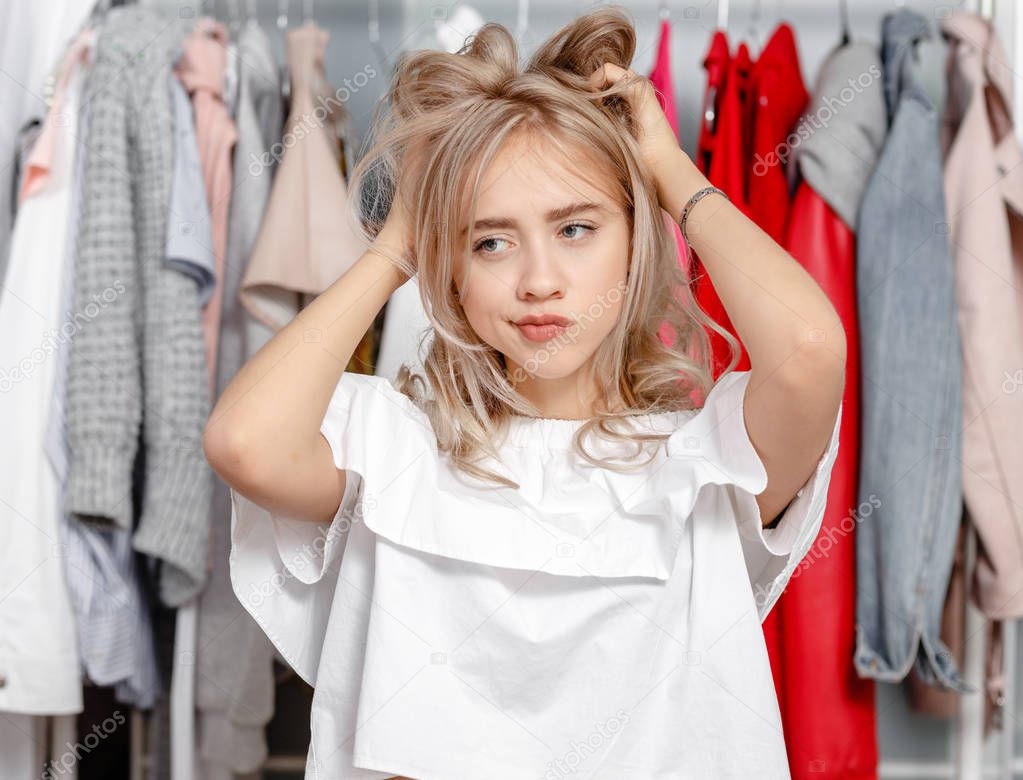 Young pretty girl blogger stands with a thoughtful expression on his face on the background of clothes hanging on a hanger in the wardrobe