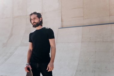 Elinde kaykay tutan şık rahat giysiler giymiş adam güneşli bir günde slayt yanında bir paten parkı duruyor