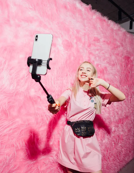 Jovem blogueiro bonita menina vestida de moda vestido rosa fazer uma selfie no fundo da parede de pele rosa na sala de show — Fotografia de Stock