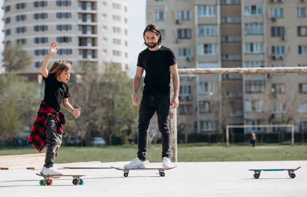 Jonge vader en zijn zoon gekleed in de stijlvolle casual kleding rijdt skateboards op een platform buiten naast het huis op de zonnige warme dag — Stockfoto