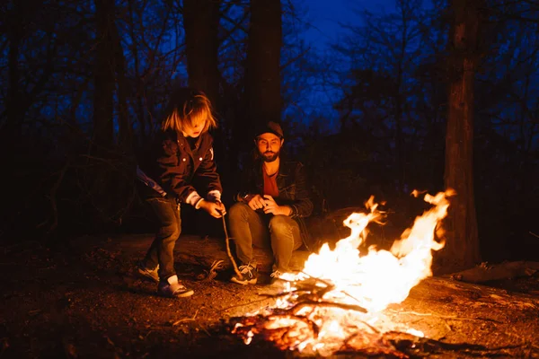 Feliz padre y su hijo sentados afuera frente a un fuego y mirando el fuego en la noche . — Foto de Stock