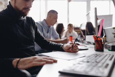 Rahat ofis tarzı giysiler giymiş adam modern ışık ofisinde meslektaşları yanında bilgisayar ile masada oturan belgeler ile çalışıyor