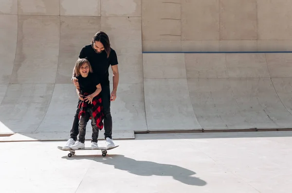 Vader en zijn kleine zoon gekleed in de casual kleding staan samen op het One skateboard in een skatepark op de zonnige dag — Stockfoto