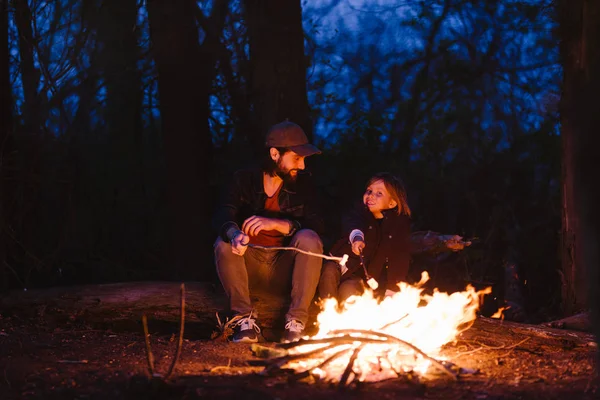 El padre y su hijo sentados en los troncos del bosque frente a un fuego y asando malvaviscos en las ramitas — Foto de Stock