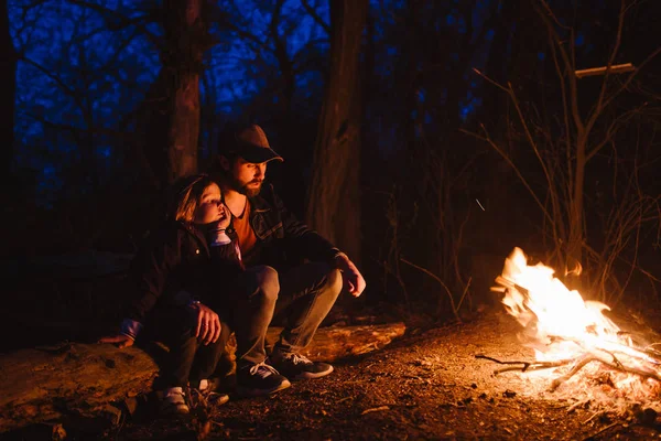 Padre y su pequeño hijo sentados juntos en los troncos frente a un fuego en la noche. La caminata en el bosque . — Foto de Stock