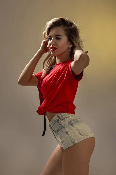 Chica rubia delgada con lápiz labial rojo vestido con pantalones cortos de mezclilla y una camiseta roja está posando contra una pared en el estudio — Foto de Stock