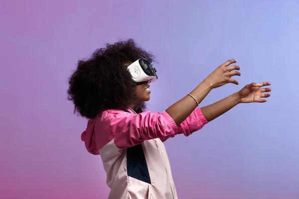 Moderna chica de pelo castaño rizado vestida con la chaqueta deportiva rosa utiliza las gafas de realidad virtual en el estudio sobre fondo de neón — Foto de Stock