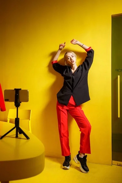 La joven bloguera de moda vestida con pantalones rojos y chaqueta negra se toma una selfie en el teléfono inteligente de pie sobre un soporte en la mesa de la habitación con paredes y muebles amarillos — Foto de Stock