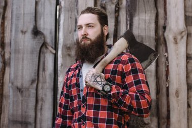 Ahşap bir çitin arka planına karşı elinde balta ile ayakta kontrol edilmiş bir gömlek giymiş sakallı acımasız güçlü adam