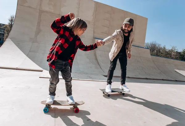 Padre y su hijo pequeño vestidos con ropa casual montan skateboards en un parque de skate con toboganes afuera en el día soleado —  Fotos de Stock