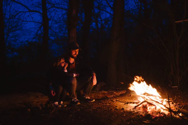 Padre y su pequeño hijo sentados juntos en los troncos frente a un fuego en la noche. La caminata en el bosque . — Foto de Stock