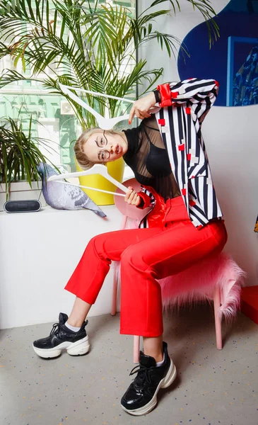 Dívčí módní blogovací sako, oblečené ve stylovém proužkované bundě a červené kalhoty, sedí na stoličce s růžovou kožešinou v místnosti na pozadí s rostlinným — Stock fotografie