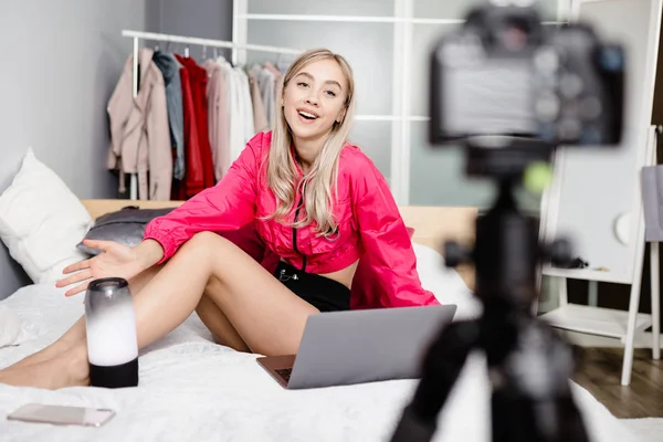 Mooie jonge meisje blogger gekleed in heldere modieuze kleding zittend op het bed met een laptop en het maken van videoclip voor reclame draagbare speakers in haar blog — Stockfoto