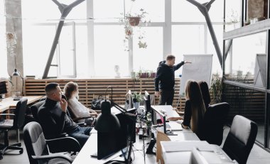 Meslektaşların toplantısı. Bir takım bilgisayarlar ve dizüstü bilgisayarlar ile masalarda oturan ve geniş bir ışık modern ofiste flipchart tarafından ayakta meslektaşım dinleme