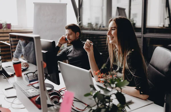 Ung brunette kledd i kontorklær jobber ved datamaskinen som sitter ved skrivebordet med kollegene på det moderne kontoret. – stockfoto