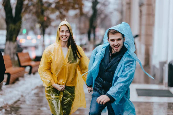 Engraçado e amoroso cara e menina em capas de chuva amarelas e azuis estão correndo na chuva lá fora . — Fotografia de Stock