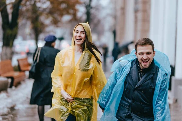 Engraçado e amoroso cara e menina em capas de chuva amarelas e azuis estão correndo na chuva lá fora . — Fotografia de Stock