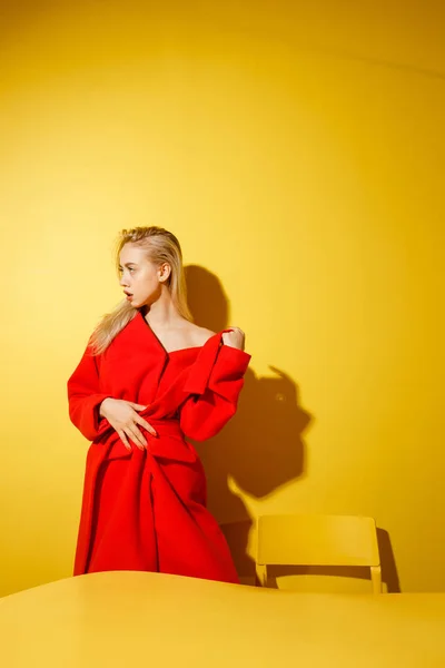 V elegantní červené kabátě stojí na žlutém stole na pozadí žlutých stěn módní blogovací dívka. — Stock fotografie