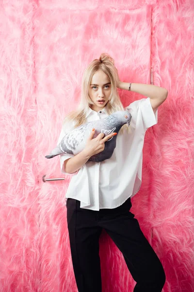 Young girl blogger dressed in stylish white shirt and black trousers poses with with a soft toy in the shape of a pigeon on the background of pink fur wall — Stock Photo, Image