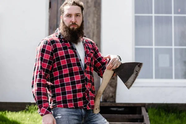 Starker brutaler Mann mit Bart und kariertem Hemd, der mit einer Axt in der Hand vor dem Hintergrund des Hauses mit großem Fenster steht — Stockfoto