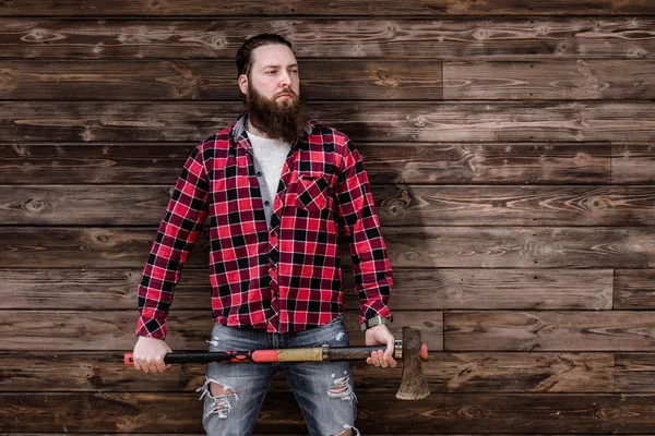 Brutal homem forte com uma barba vestida com uma camisa marcada e jeans rasgado fica com um machado nas mãos no fundo de uma parede de madeira — Fotografia de Stock