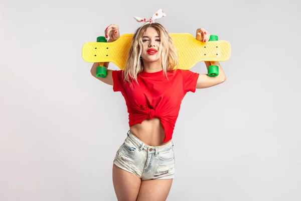 Jovem vestida com shorts curtos de ganga e uma camiseta vermelha posa com um skate amarelo no estúdio — Fotografia de Stock