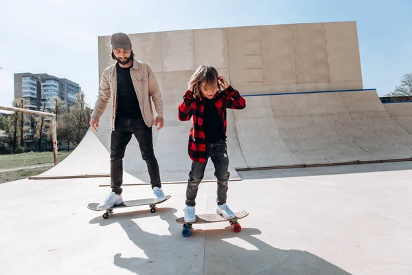 Vader en zijn kleine zoon gekleed in de casual kleren Ride Skateboards in een skatepark met glijbanen buiten op de zonnige dag — Stockfoto