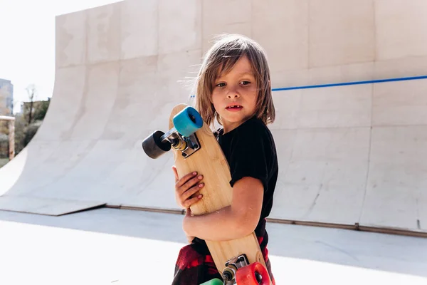 De jongen gekleed in de casual kleding met skateboard in zijn hand staat in een skatepark naast de glijbaan op de zonnige dag buiten — Stockfoto