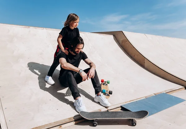 Glücklicher junger Vater und sein Sohn in stylischer Freizeitkleidung sitzen bei sonnigem warmen Tag in einem Skatepark in einer Umarmung zusammen auf der Rutsche neben den Skateboards — Stockfoto