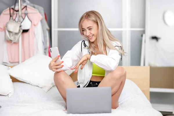 Посміхаючись красива дівчина блогер одягнений в модний одяг бере селфі на свій смартфон для її блог, сидячи на ліжку з ноутбуком в кімнаті — стокове фото
